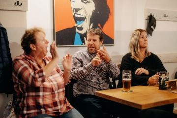 Dvě ženy a jeden muž sedí u stolu. Žena s mužem spolu komunikují ve znakovém jazyce.