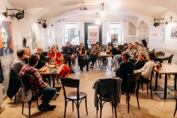 Místnost plná lidí, kteří sedí v kruhu na židlích. Všichni se dívají na jednu ženu, která komunikuje ve znakovém jazyce.