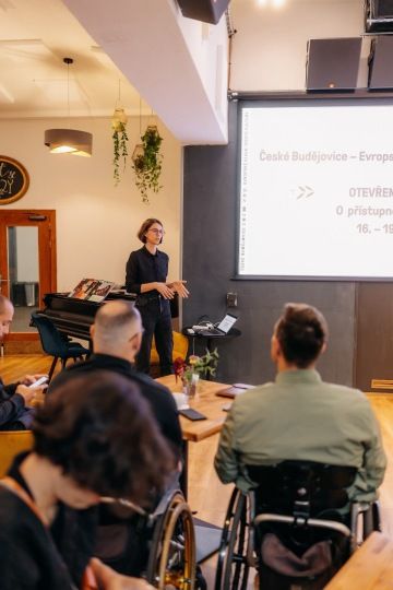 Lidí se dívají na prezentaci s motivy Evropského hlavního města kultury. Žena stojí vedle promítacího plátna s prezentací a povídá.