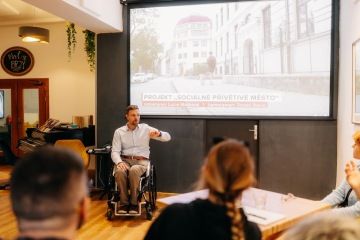 Diváci se dívají na plátno s prezentací, kde je fotka ulice s titulkem “sociálně přívětivé město”. U prezentačního plátna je muž na vozíku a mluví.