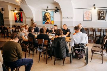 Skupinové setkání u kulatého stolu v kavárně, lidé diskutují o přístupnosti a programu EHMK 2028, v pozadí visí pop-artové portréty.