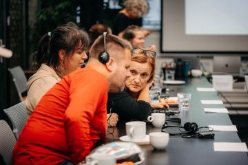 Sedící muž se sluchátky se naklání a hovoří k ženě, která sedí vedle jeho sousedky u společného stolu.