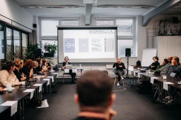 Lidé se dívají na prezentaci s motivy Evropského hlavního města kultury. Komentuje ji žena sedící nalevo, vpravo překladatelka do znakového jazyka.