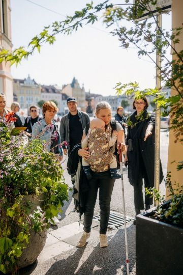 Skupina lidí prochází městem, žena s bílou holí a dítětem v nosítku jde v čele, v pozadí hlavní náměstí Českých Budějovic.