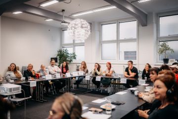 Účastníci setkální Aliance pro přístupnost sedí kolem velkého stolu do U který vyplňuje celou místnost, na stole jmenovky a pracovní materiály.