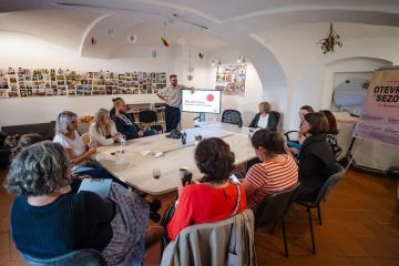 Skupina lidí sedí kolem stolu a dívají se na prezentaci v televizi. Vedle televize stojí muže, který se směje.