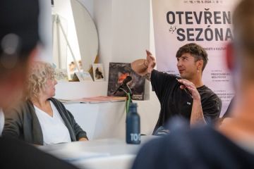 Muž a žena sedí u stolu. Muž něco popisuje a gestikuluje, žena se na něj dívá.