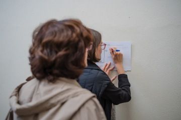 Dvě ženy zezadu. Jedna žena stojí u zdi, o kterou má opředený papír a píše.