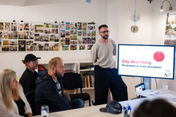 Muž prezentuje lidem kolem stolu projekt „City describing – the practice of audio description“, na obrazovce je název programu, v místnosti visí fotografie z komunitních aktivit.
