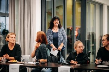 Ve spodní části sedí za stolem 4 ženy, jedna z nich je na vozíku. Nad nimi je v záběru další žena, která právě vešla do dveří.